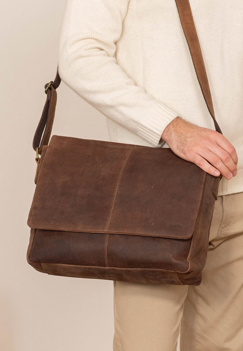 Brązowa skórzana torba na ramię z płaskim klapkiem, regulowanym paskiem i widocznym przeszyciem. Gładka faktura o lekko zużytym wyglądzie.