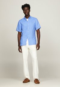 Camicia di lino azzurro chiaro a maniche corte con colletto polo, abbinata a pantaloni bianchi e scarpe marroni. Design semplice, linee pulite, vestibilità casual.