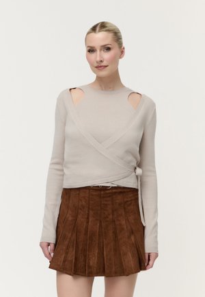 Femme aux cheveux blonds portant un pull beige avec découpes et une jupe plissée en daim marron, debout devant un fond uni clair.