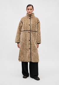 Viltota kažokāda gaiši brūnā krāsā ar teksturētu rakstu, melniem apmalēm, augstu apkakli un priekšējiem pogu aizdariem. Modelis valkā melnas platās bikses.