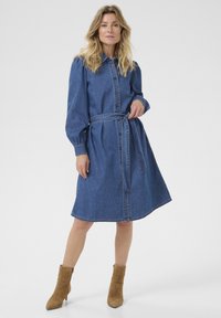 Denim mekko, sininen, nappikiinnitys edessä, pitkät puff-hihat ja vyötäröllä vyö. Pehmeä tekstiili ja polven pituinen helma. Yhdistetty vaaleanruskeisiin nilkkureihin.