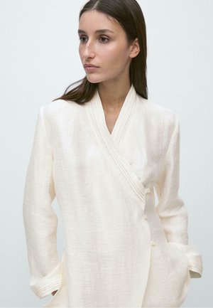 Kobieta o długich brązowych włosach, ubrana w kremową teksturowaną kurtkę typu wrap, z rękami w kieszeniach, patrząca na bok na jednolitym tle.