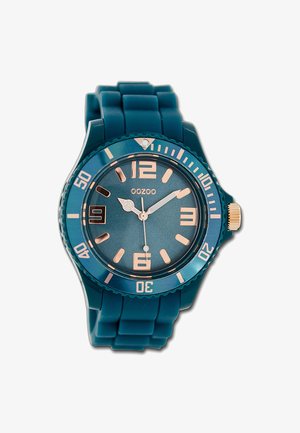 Montre en silicone teal avec un cadran rond, des accents en or rose, des marquages numériques audacieux et une lunette texturée. Comprend une fenêtre de date à 6 heures.