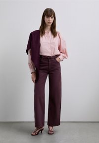 Lichtroze blouse met lange mouwen en knoopsluiting, met geplooide ballonmouwen, gecombineerd met hooggetailleerde, brede donderpaarse broekspijpen en sandalen met bandjes.