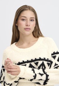 Maglione crema realizzato in tessuto a maglia testurizzato, con un audace motivo geometrico nero lungo le maniche e il torace, caratterizzato da un ampio scollo.