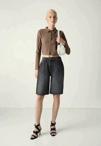 Brūns rievojums garām piedurknēm polo krekls, augstā vidukļa tumšas džinsu bermudu šorti, melnas siksniņu papēžu kurpes un baltā rokassoma. Vienkārša fona.