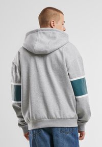Haut à capuche gris avec une grande capuche, présentant des accents rayés turquoise et blanc sur les manches. Fabriqué en tissu doux, avec des poignets et un ourlet côtelés.