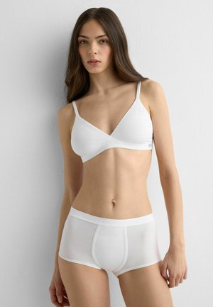 Jonge vrouw met lang bruin haar, gekleed in een witte bralette en witte short-style onderbroek, staand tegen een witte muur.