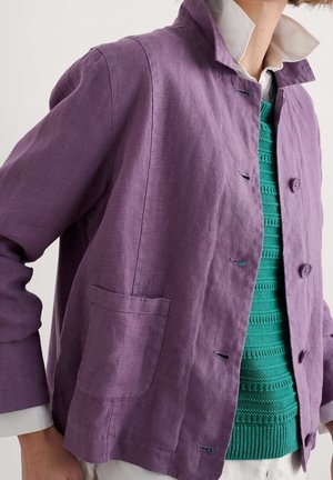 Personne portant une veste en lin violette boutonnée par-dessus un pull vert texturé et une chemise blanche à col.