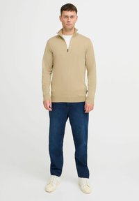 Beige strikkegenser med halvglidelås i kragen, kombinert med mørkeblå jeans og lyse sportsko. Modellen står mot en ensfarget bakgrunn.