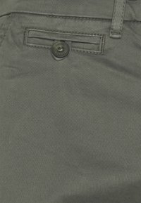 Donkergroene stoffen broek met een gladde textuur, voorzien van een voorzak afgesloten met een zwarte knoop en gestikte accenten.