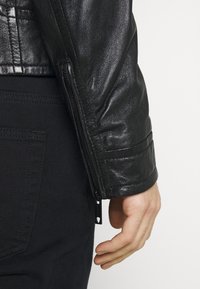 Zwarte leren jas met een gestructureerde afwerking, voorzien van een ritssluiting aan de mouw en subtiele stiksel details, gecombineerd met donkere stoffen broek.