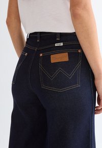 Rear view of person wearing dark blue Wrangler jeans with visible back pockets and brand label, paired with a white shirt.