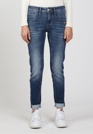 Femme portant un jean slim bleu retroussé avec poches avant et baskets blanches Nike, debout devant un fond uni.