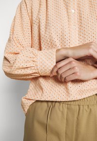 Frau in einer Pfirsichfarbenen Bluse mit Lochmuster und puffierten Ärmeln, die beigen hochgeschnittenen Hosen trägt, mit sanft verschränkten Händen an der Taille.