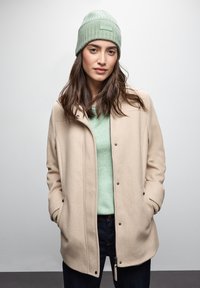 Cappotto beige con bottoni a pressione, polsini a coste e una finitura testurizzata, indossato sopra un maglione verde chiaro e abbinato a un berretto in maglia verde menta.