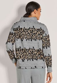 Frau mit dunklem Haar in einem tiefen Pferdeschwanz, die einen grauen Pullover mit braunen und schwarzen Leopardenmusterstreifen trägt und von hinten schaut.