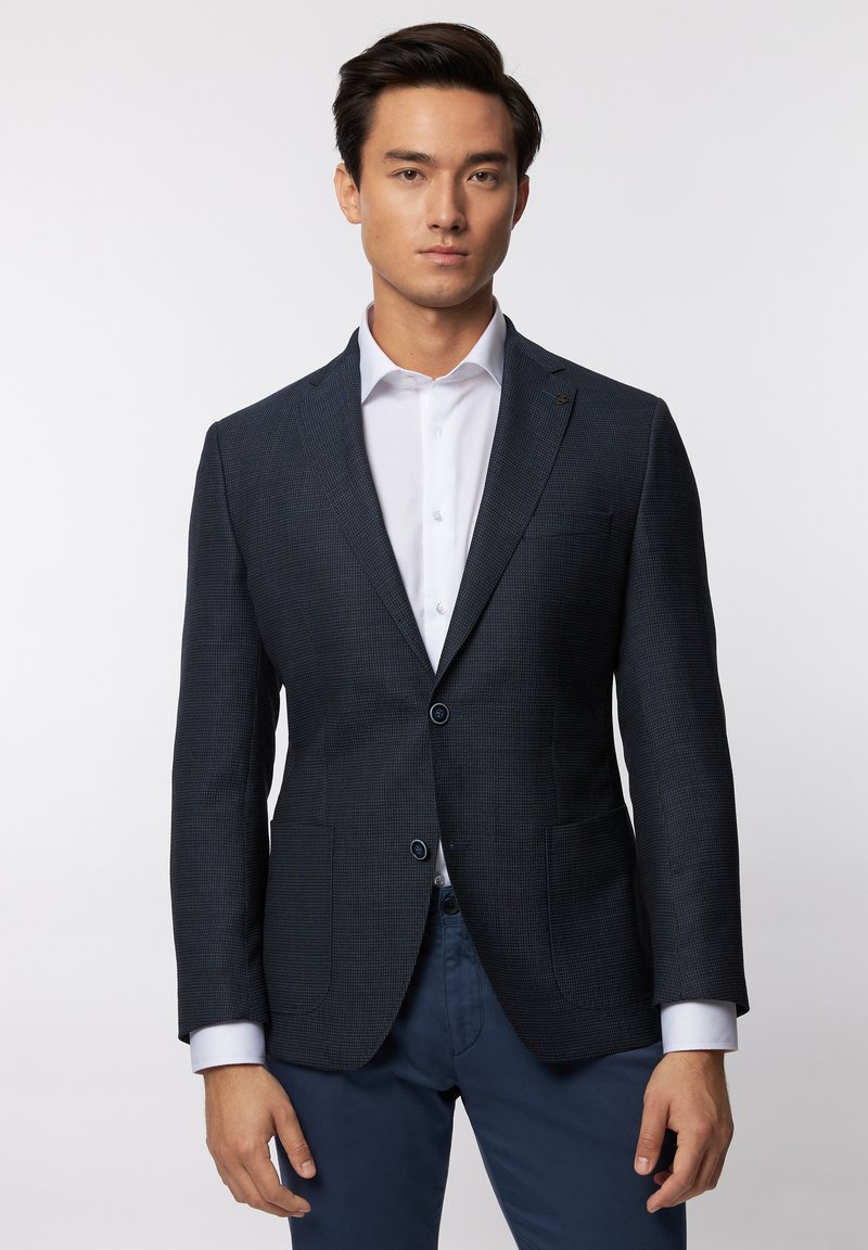 Blazer texturé marine avec deux poches avant et deux boutons, associé à une chemise blanche et un pantalon bleu, offrant une coupe moderne et ajustée.