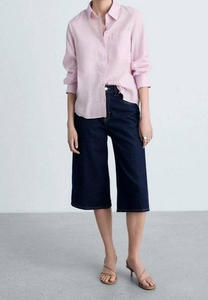 Donna che indossa una camicia rosa chiaro con bottoni, pantaloncini di jeans larghi scuri fino al ginocchio e sandali beige con tacco e cinturini.