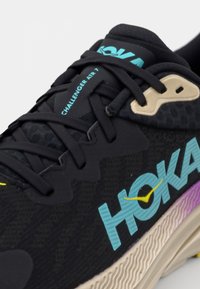 Baskets de sport noires avec tige en mesh, présentant un logo turquoise, des accents crème, et une semelle texturée passant du beige au violet.