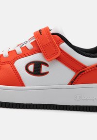 White and orange sneaker with a textured finish, featuring a Velcro strap, black accents, and a prominent logo on the side. Rubber sole.