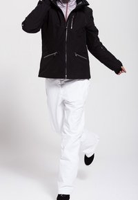 Svart vattentät jacka med dragkedjefickor och justerbar huva, tillsammans med vita snöbyxor och svart fotbeklädnad, som visar en stilren design.
