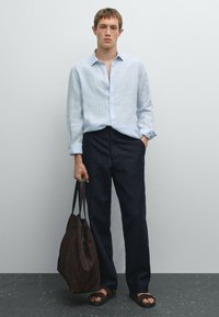 Camicia in lino azzurro chiaro con bottoni e maniche lunghe, abbinata a pantaloni scuri a righe. Borsa a tracolla in suede marrone e sandali scuri.