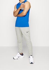 Hombre con una camiseta de tirantes atlética azul, pantalones de chándal grises de Nike y zapatillas blancas de Nike sostiene una botella de agua contra un fondo blanco liso.