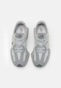 Pár světle šedých tenisek New Balance s detailním prodyšným materiálem a semišem, šněrovacím uzávěrem a polstrovaným límcem, pozorováno shora na bílém pozadí.