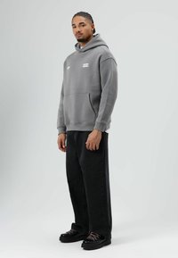 Grijze hooded sweater met een voorvak en branding; gecombineerd met zwarte loszittende jeans en zwarte schoenen met roze veters.