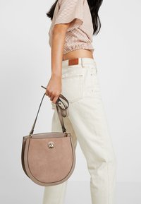 Borsa rotonda di colore beige-rosato realizzata in suede con una finitura liscia; presenta un accessorio in metallo circolare dorato e una tracolla staccabile.