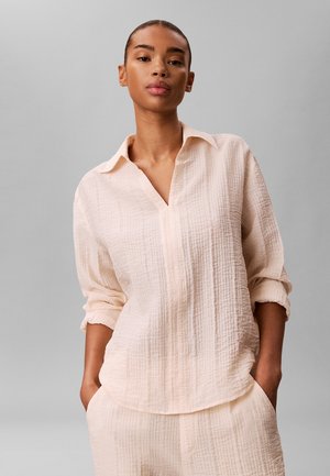 Donna in piedi con le mani nelle tasche, indossa una camicia a maniche lunghe rosa chiaro con trama e pantaloni abbinati su uno sfondo grigio.