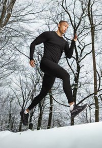 Zwarte sportieve langetermijn shirt en leggings gedragen door een hardloper in de sneeuw. Kenmerken zijn een gestroomlijnde fit en lichtgewicht materiaal. Zwarte hardloopschoenen.