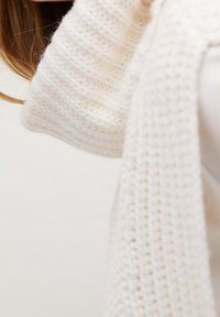 Bílý pletený svetr s ribovým texturováním, který se vyznačuje měkkým a robustním designem. Látka vypadá silně a útulně s volným střihem.