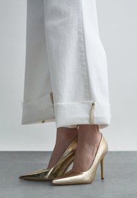 Personne portant un pantalon blanc à jambes larges avec des ourlets bruts et des escarpins métalliques dorés à bouts pointus, debout sur un sol gris.