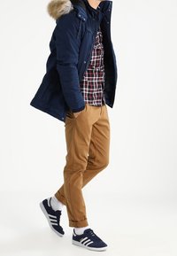 Mörkblå vadderad jacka med pälshuva, rutmönstrad skjorta, beige byxor och mörkblå sneakers med vita ränder. Avslappnad outfit på en modell i rörelse.