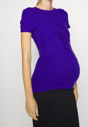 Lilla kortærmet top med ribstrik og rynket design samt justerbar snøre foran, båret over en sort nederdel, der fremhæver en gravid silhuet.