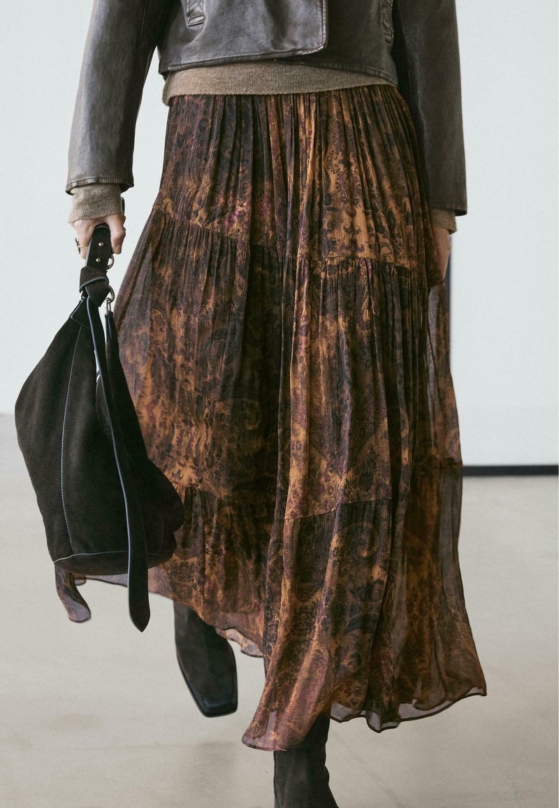 Femme portant une jupe longue marron transparente à motifs, une veste en cuir et des bottes noires, tenant un sac à main en daim noir dans une main.