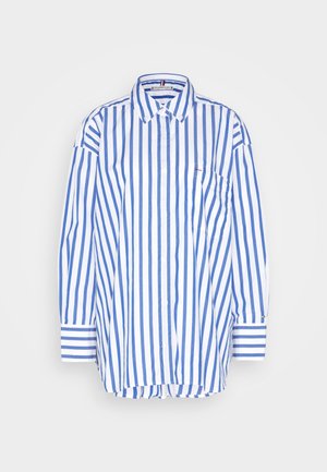 Sinivalkoinen pystysuunnassa raidallinen pitkähihainen napitettava paita, jossa on etutasku rinnassa ja napillinen kaulus, vaaleanharmaalla taustalla.