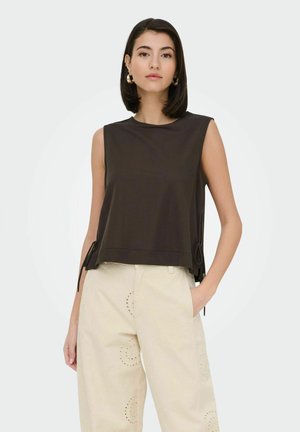 Femme aux cheveux foncés jusqu'aux épaules porte un haut noir sans manches avec liens sur les côtés et un pantalon crème taille haute avec motifs circulaires cousus.