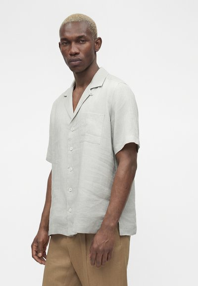 Άνδρας με κοντά ξανθά μαλλιά, φοράει ανοιχτό γκρι κοντομάνικο πουκάμισο με κουμπιά και μπεζ παντελόνι, στέκεται μπροστά σε απλό φόντο.