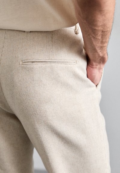 Man wearing beige textured pants and beige shirt, hand in front pocket, showing back and side detail with welt pocket.