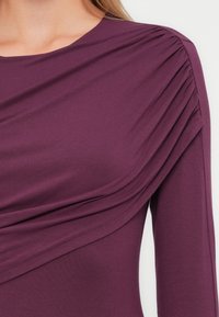 Haut violet profond avec des détails froncés à l'avant et des manches longues, fabriqué à partir d'un tissu lisse et extensible avec un léger éclat.