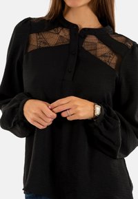 Blusa nera con tessuto strutturato, yoke in pizzo trasparente, chiusura frontale con bottoni e maniche a sbuffo. Accentuata da una sottile lucentezza e design aderente.