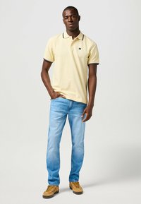 Camisa polo de color amarillo pálido con ribete negro en el cuello, combinada con unos jeans azul claro y zapatos marrones. La camisa presenta un pequeño logo en el pecho.