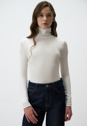 Weißes, geripptes Turtleneck-Oberteil mit langen Ärmeln, kombiniert mit hochgeschnittenen, dunkelblauen Jeans aus Denim mit Knopfverschluss und orangefarbenen Nähten.