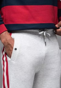 Graue Jogginghosen mit roten gestreiften Seitenakzenten, elastischem Bund mit Kordelzug und Seitentaschen mit schwarzen Druckknöpfen.