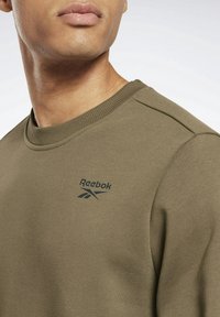 Světle olivová mikina vyrobená z měkké tkaniny s žebrovaným výstřihem. Na hrudi se nachází černé logo Reebok a má jednoduchý design.