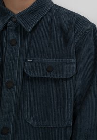 Tmavě modrá kordurová košilová bunda s límcem, dvěma předními kapsami s klopy a černými patentními knoflíky. Textura je ribbovaná a měkká.