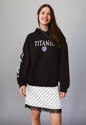 Mlada ženska se smehlja, oblečena v črno »TITANIC« kapucar s srčnim vzorcem in belo krilo s črnimi pikicami ter črnim čipkastim robom, na preprostem sivem ozadju.